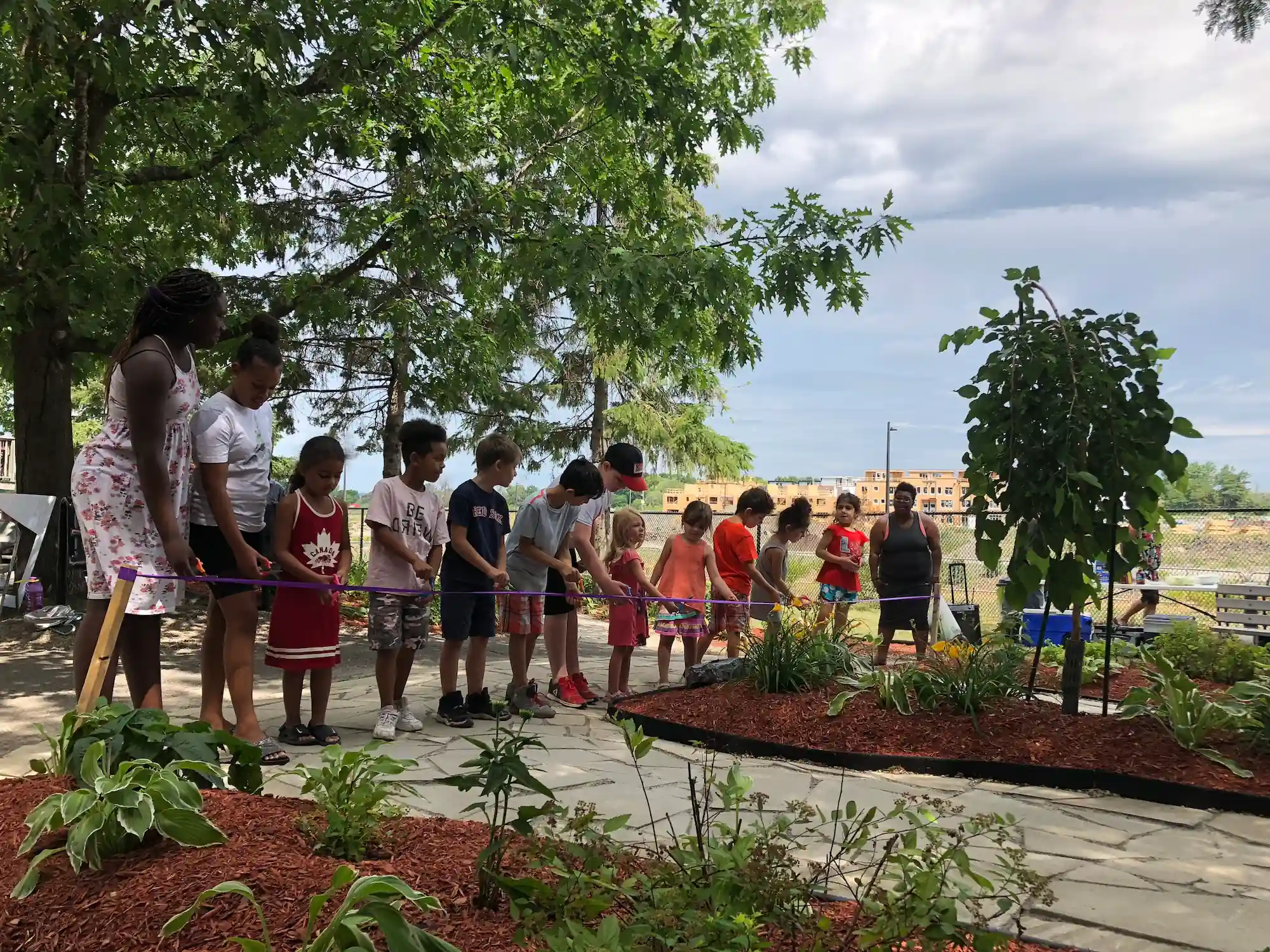 A group of children and an adult stand in a line outside, holding a ribbon for a ceremony in a co-operative housing garden with plants and trees under a cloudy sky.