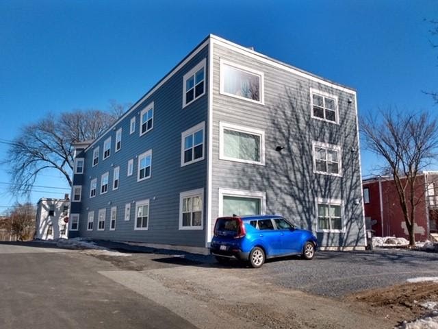 A three-story gray co-operative housing apartment building with multiple white-trimmed windows, a blue car parked in front, and leafless trees nearby under a clear blue sky.