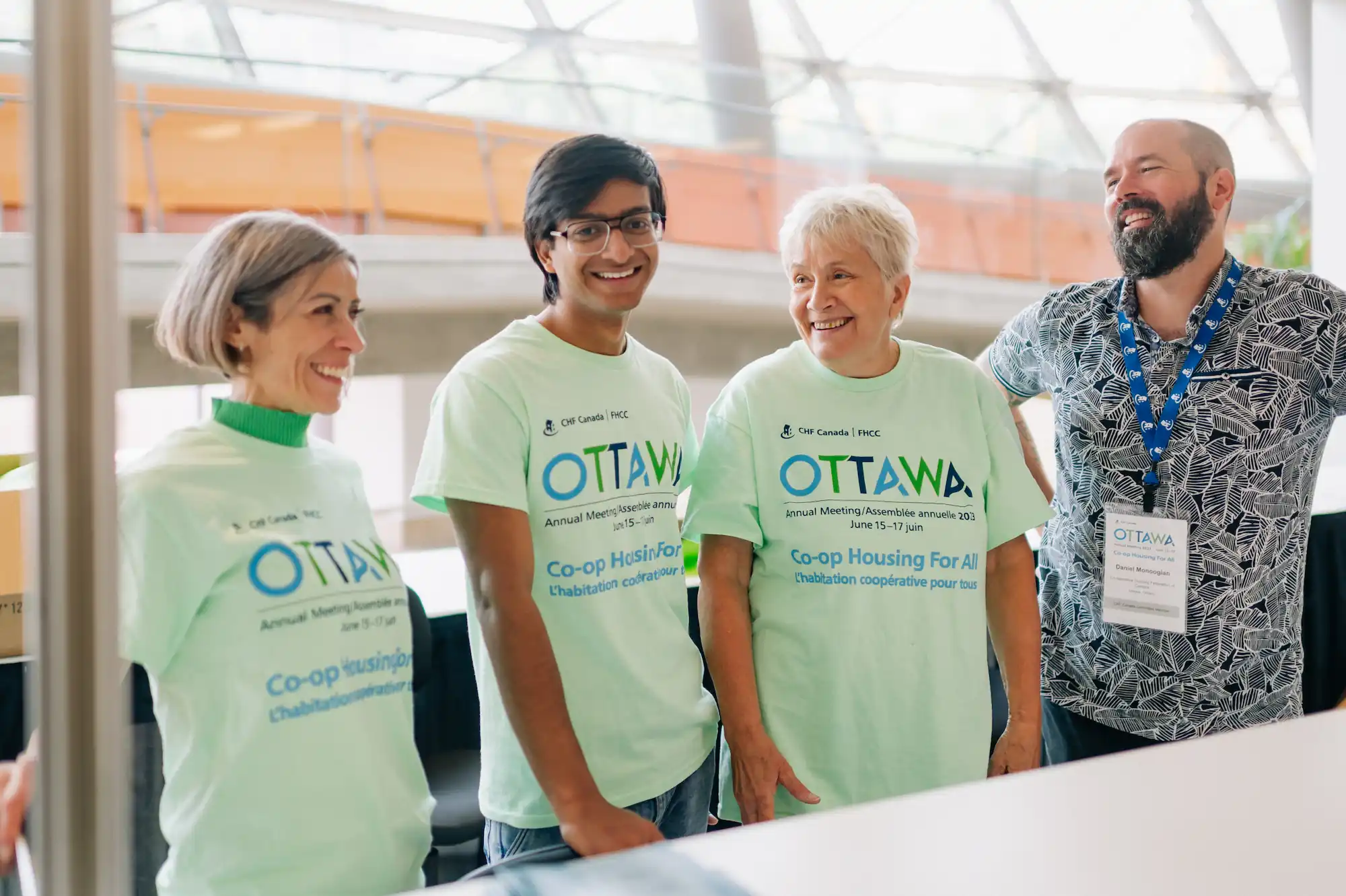 Four people stand together indoors, smiling and wearing light green "Ottawa Co-op Housing for All" t-shirts at a CHF Canada conference event, supporting the vision of Co-operative Housing.