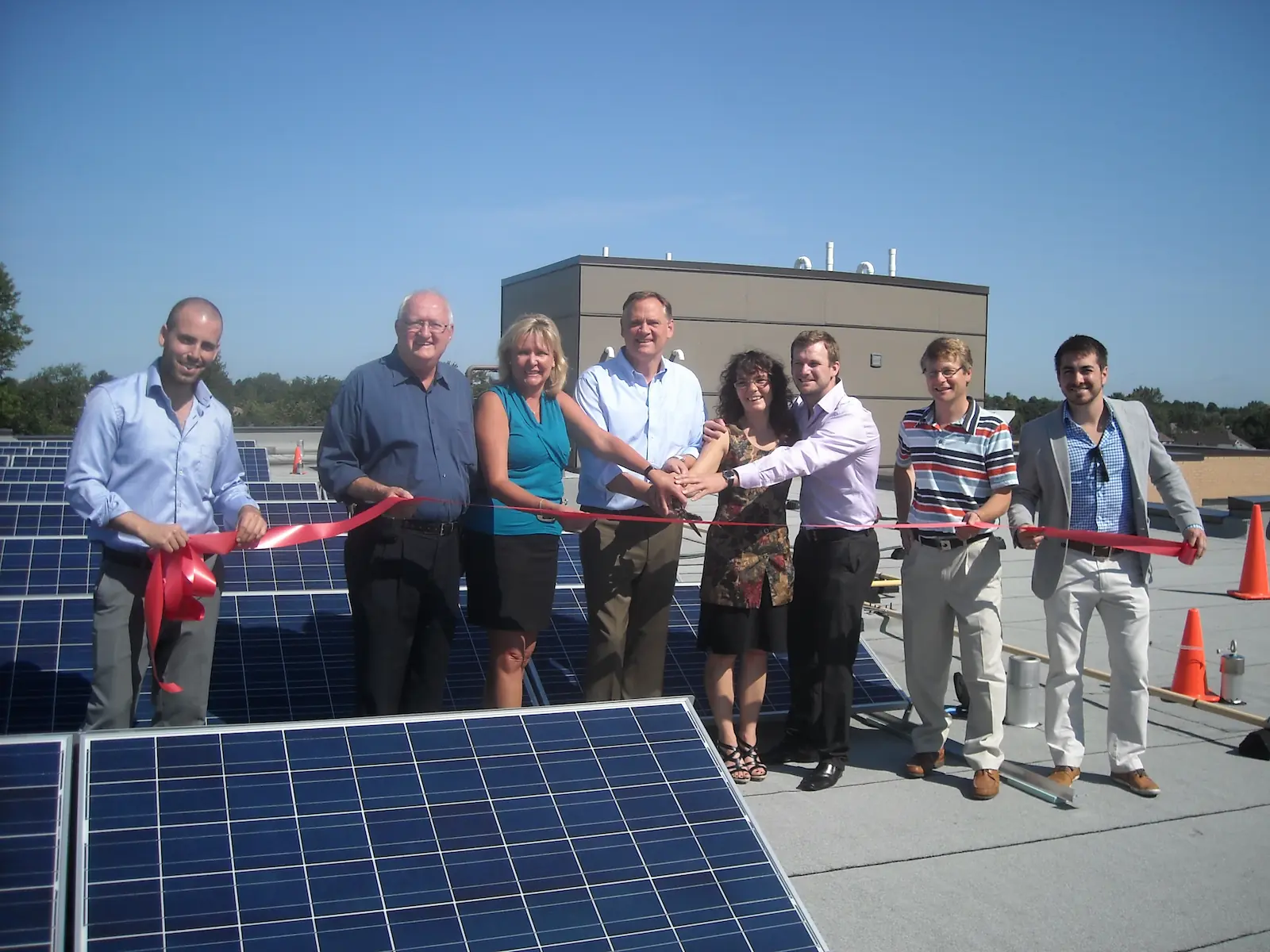 A group of people standing on a rooftop among solar panels, holding a red ribbon for a ribbon-cutting ceremony under a clear sky, celebrating the launch of new co-operative housing with CHF Canada.