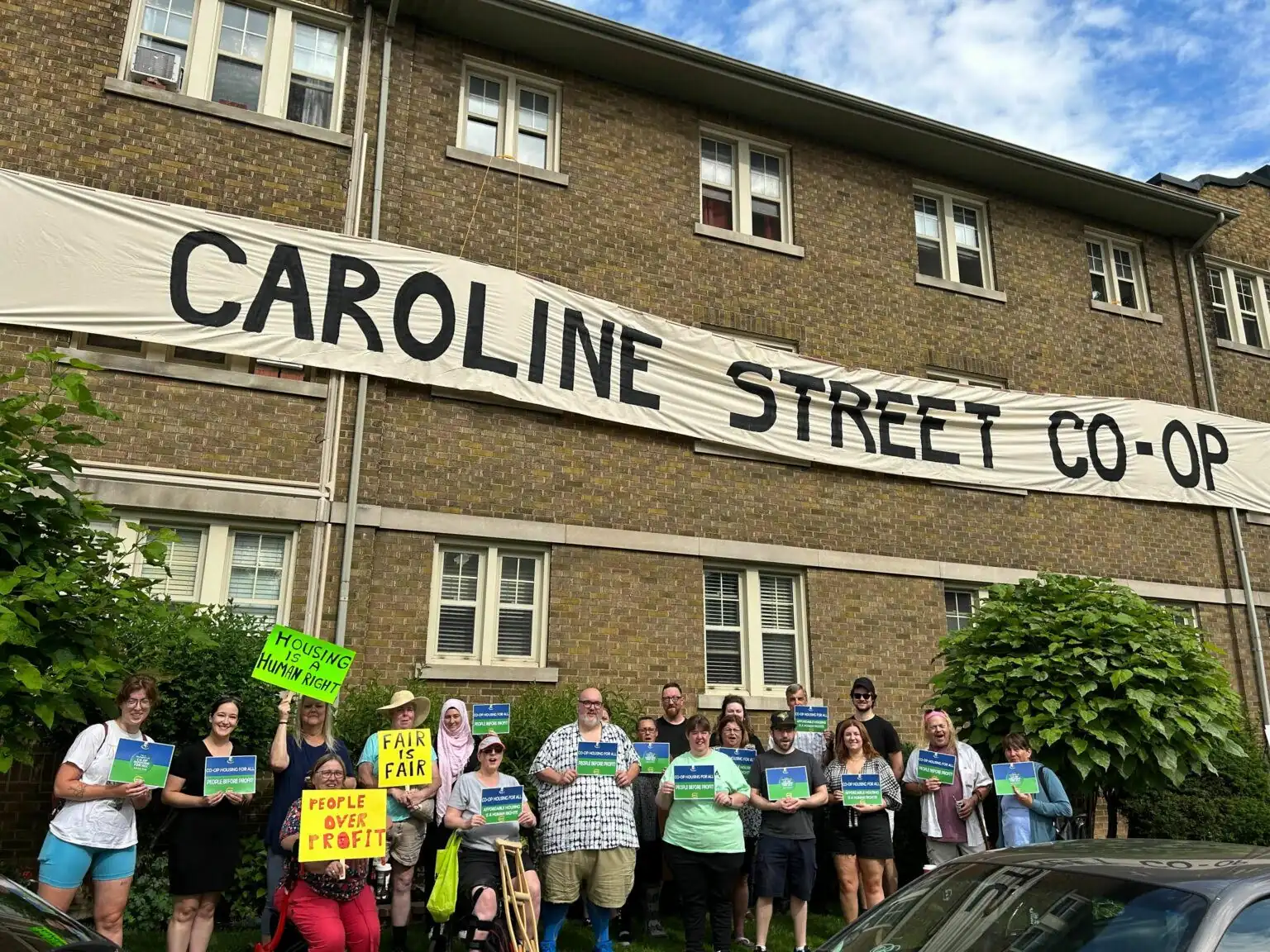 A group of people stand in front of a brick building with a large "Caroline Street Co-op" banner, holding signs and papers during a housing protest supporting Co-operative Housing and organizations like CHF Canada.