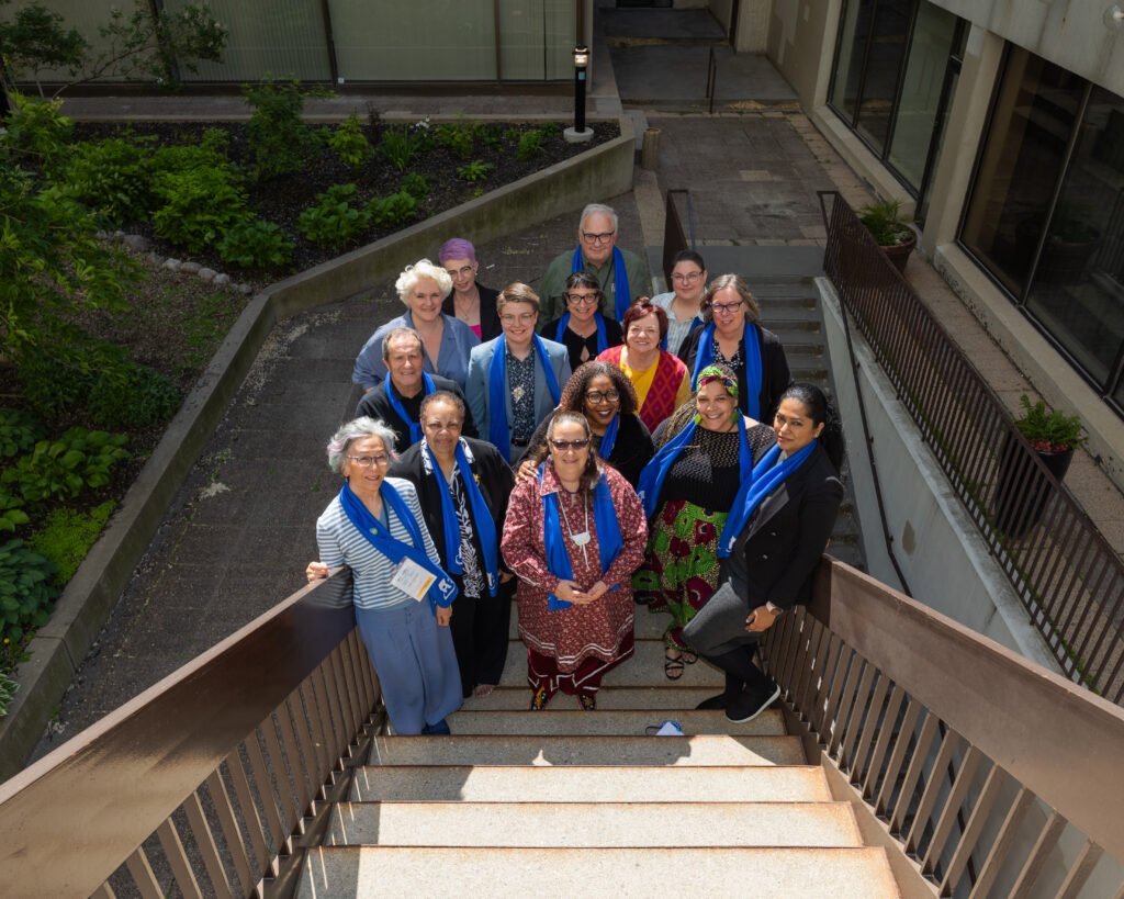 A group of sixteen adults, wearing blue scarves, stand together on outdoor stairs and a landing in a courtyard, posing for a group photo to celebrate Co-operative Housing with CHF Canada.