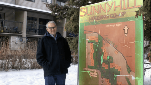 An older man in a dark coat stands outside in the snow next to a large Sunnyhill Housing Co-op map sign, representing the spirit of co-operative housing with buildings and trees visible in the background.