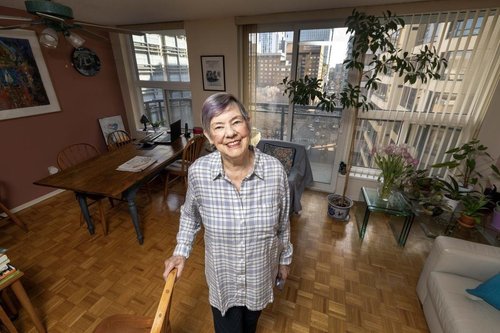 An older woman with short gray hair stands smiling in a bright co-operative housing apartment with large windows, plants, and wooden furniture.