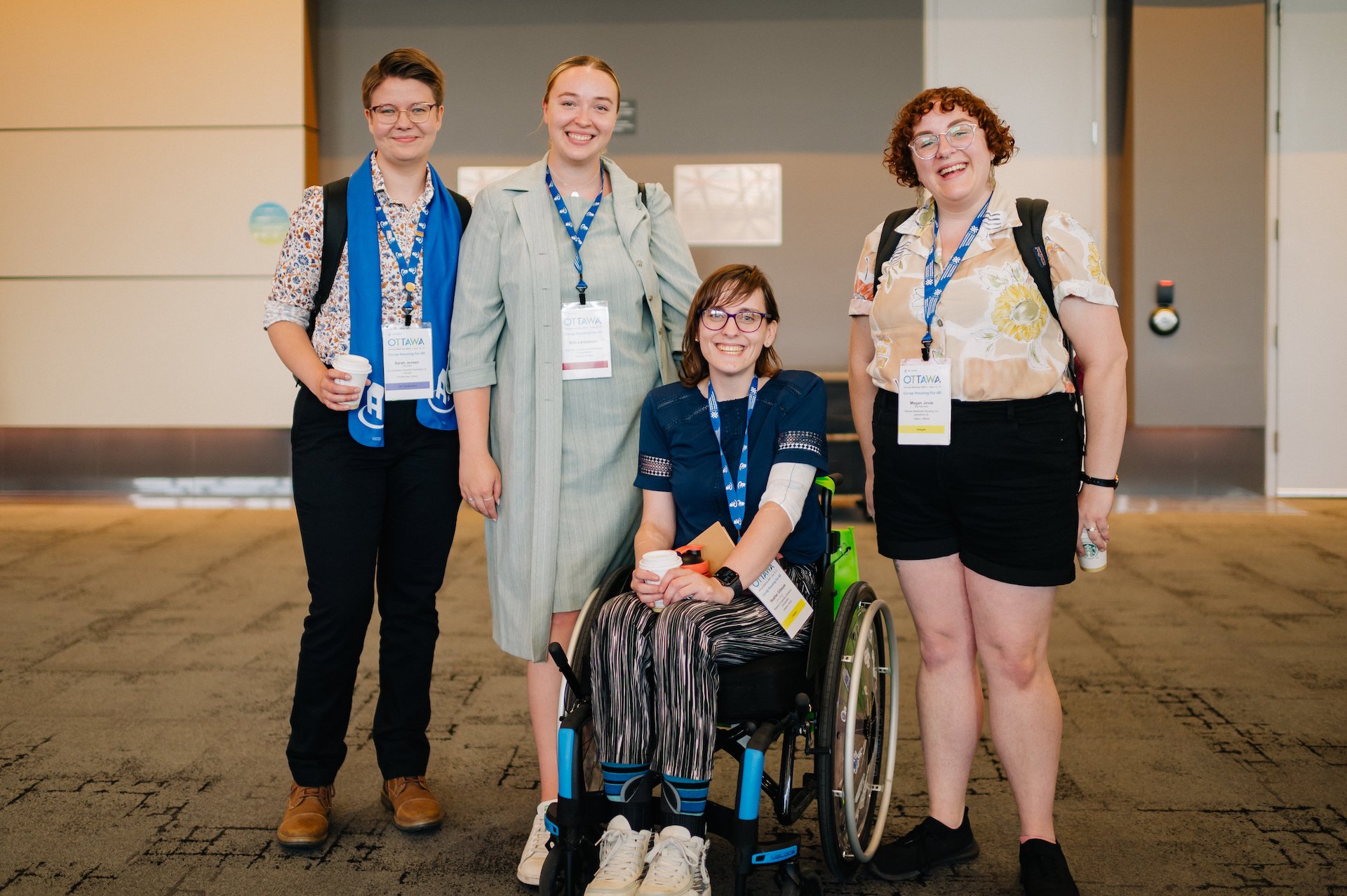Four people, three standing and one seated in a wheelchair, pose together indoors wearing CHF Canada conference badges and lanyards—united by their passion for co-operative housing.
