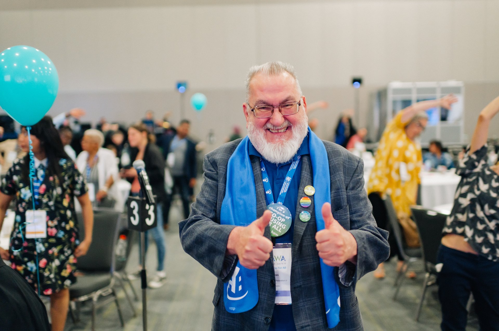 An older man with a beard smiles and gives two thumbs up at an indoor Co-operative Housing event, wearing a blue scarf and several buttons. People and tables are visible in the background.