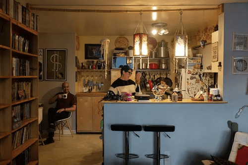 Three people are in a cozy co-operative housing kitchen; one sits in the corner holding a mug while two stand by the counter amid shelves, appliances, and hanging decorations—a warm scene inspired by CHF Canada.