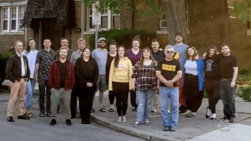 A group of 17 adults stand together on a sidewalk in a residential neighborhood, posing for a photo in daylight to celebrate their co-operative housing community.