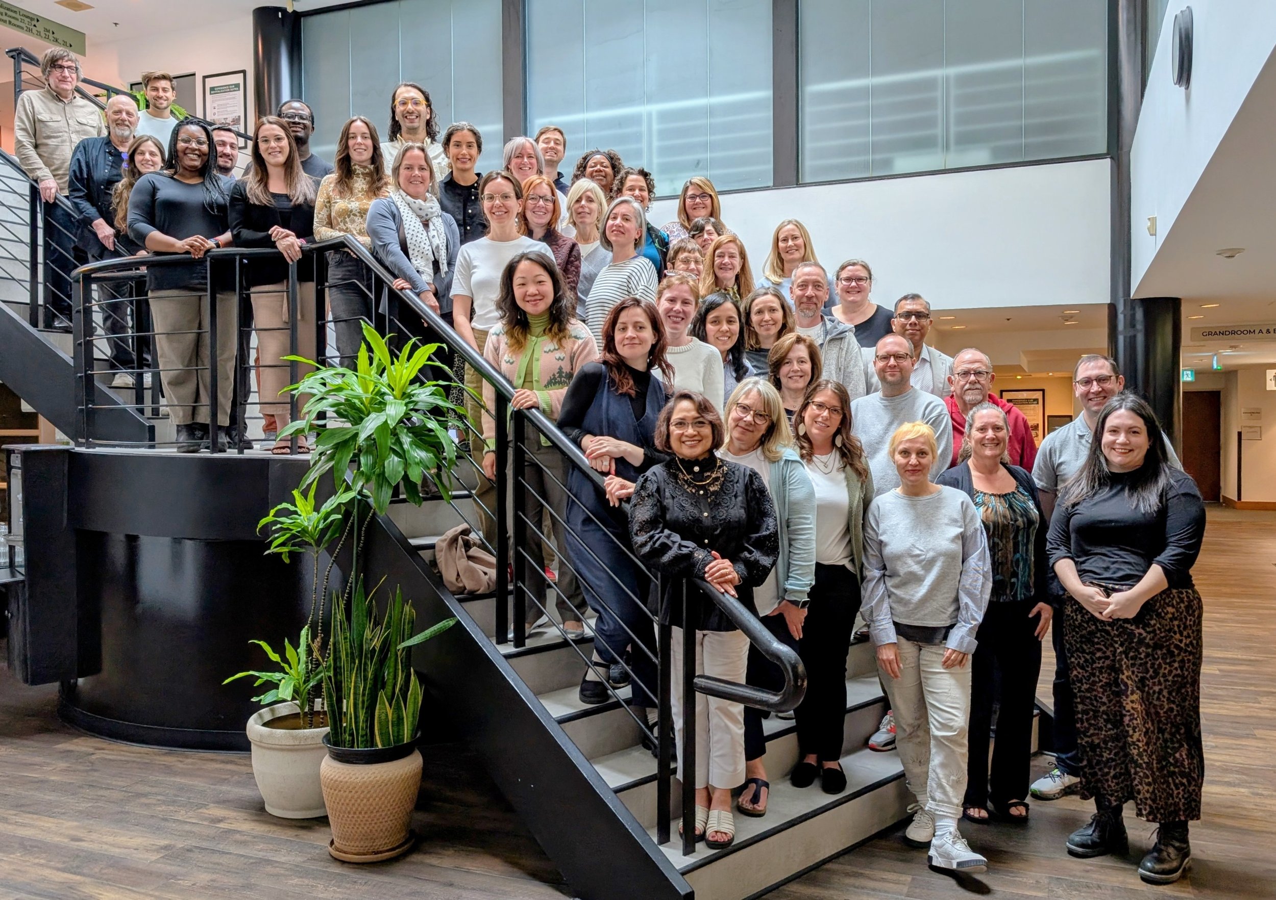 A large group of people poses together on a staircase in a modern indoor setting, smiling at the camera. There are potted plants nearby, highlighting the welcoming spirit of co-operative housing with CHF Canada.