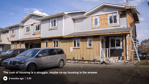 Two-story co-operative housing townhouse complex under renovation with exposed wood and construction materials, parked vehicles in front, and a cloudy sky overhead.