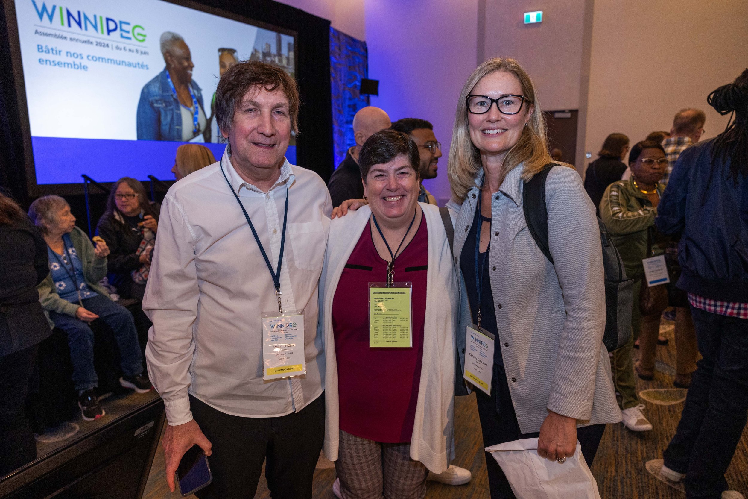 Three people stand together and smile at a Co-operative Housing conference event, with others seated in the background and a presentation screen displaying "WINNIPEG" and French text, highlighting CHF Canada’s commitment to housing.
