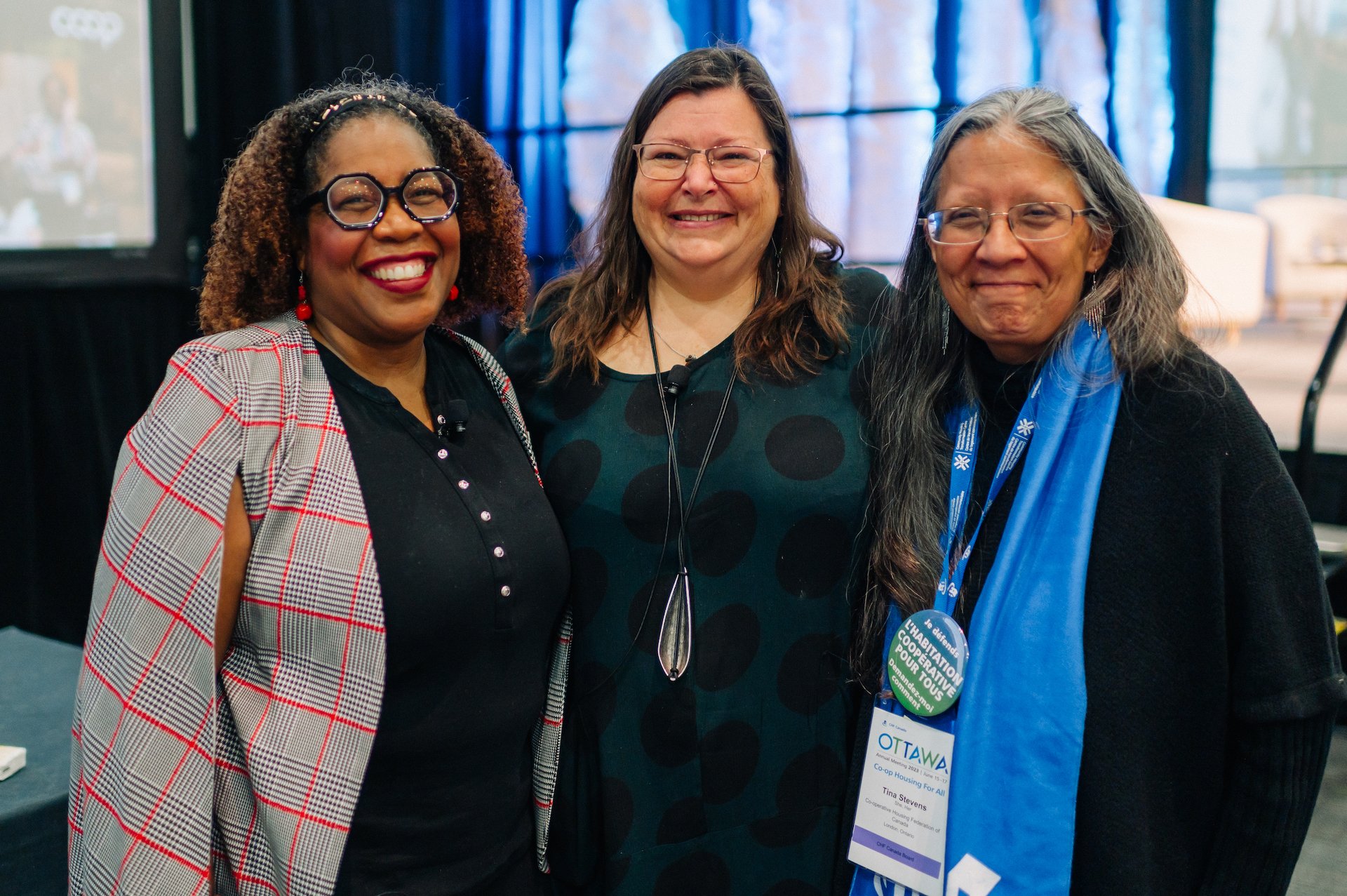 Three women stand together indoors at a CHF Canada event, smiling at the camera; one woman wears event badges and a blue scarf, highlighting their shared commitment to co-operative housing.