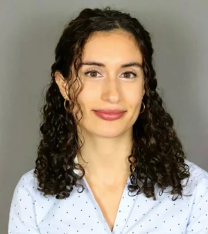 Woman with curly dark hair, wearing a light blue shirt with small dark dots, smiles slightly in front of a plain gray background, embodying the welcoming spirit often seen in Co-operative Housing communities supported by CHF Canada.