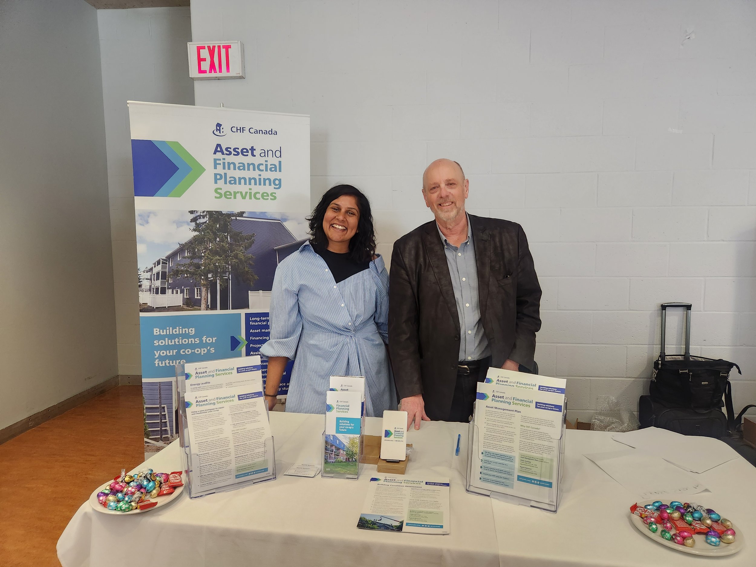 Two people stand behind a table with brochures and chocolate at CHF Canada’s Asset and Financial Planning Services booth. A banner and handouts promote Co-operative Housing initiatives to interested visitors.