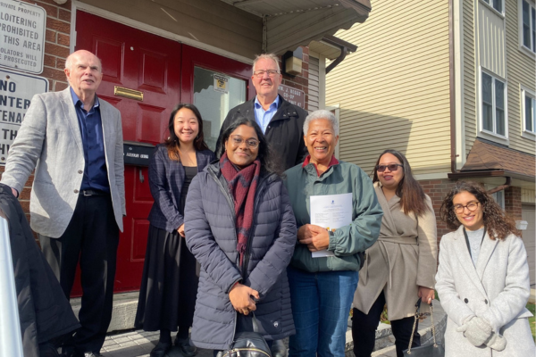 A group of seven adults poses and smiles for a photo outside a co-operative housing building with a red door on a sunny day.