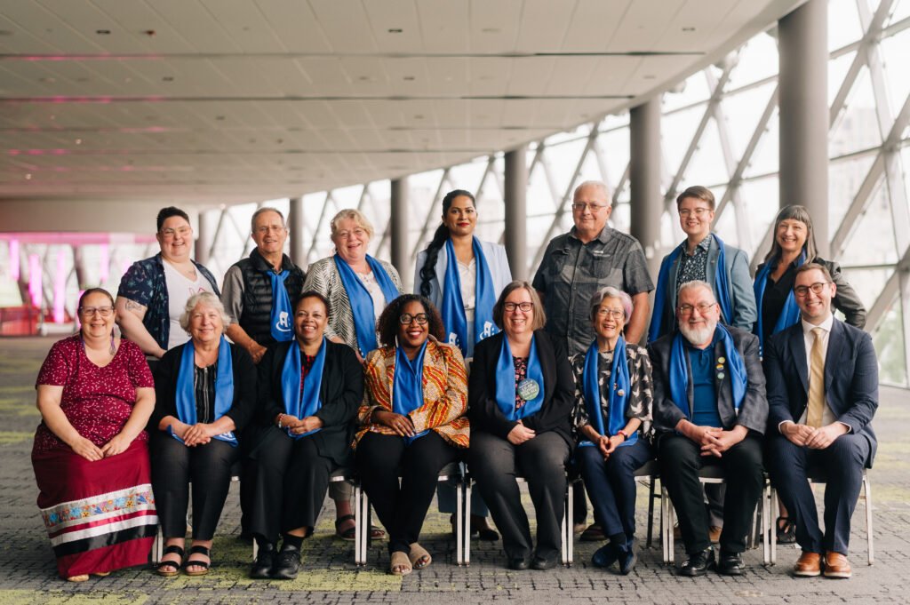 A group of fifteen people, some seated and some standing, pose indoors wearing blue scarves or sashes. Large windows line the background, highlighting their commitment to Co-operative Housing and CHF Canada.