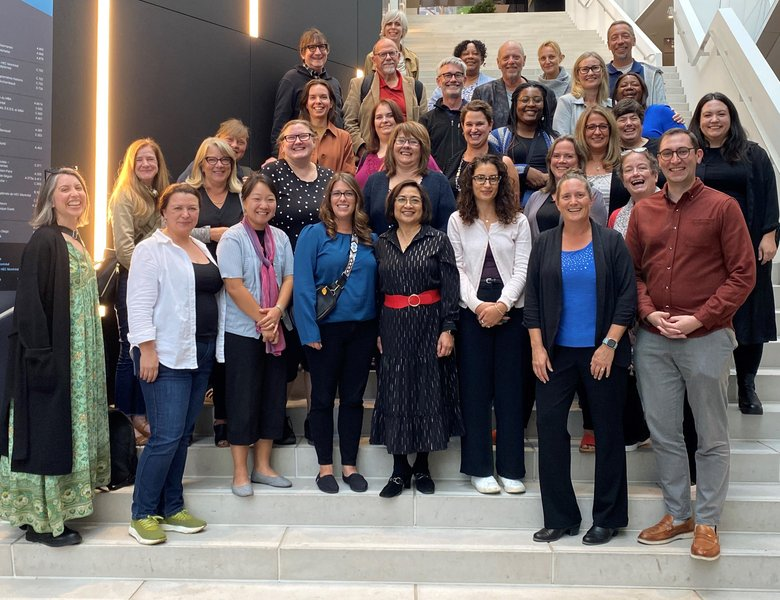 A group of about 30 adults poses together on indoor steps, smiling at the camera. The modern setting with bright lighting and a digital display in the background suggests a gathering related to Co-operative Housing or CHF Canada.