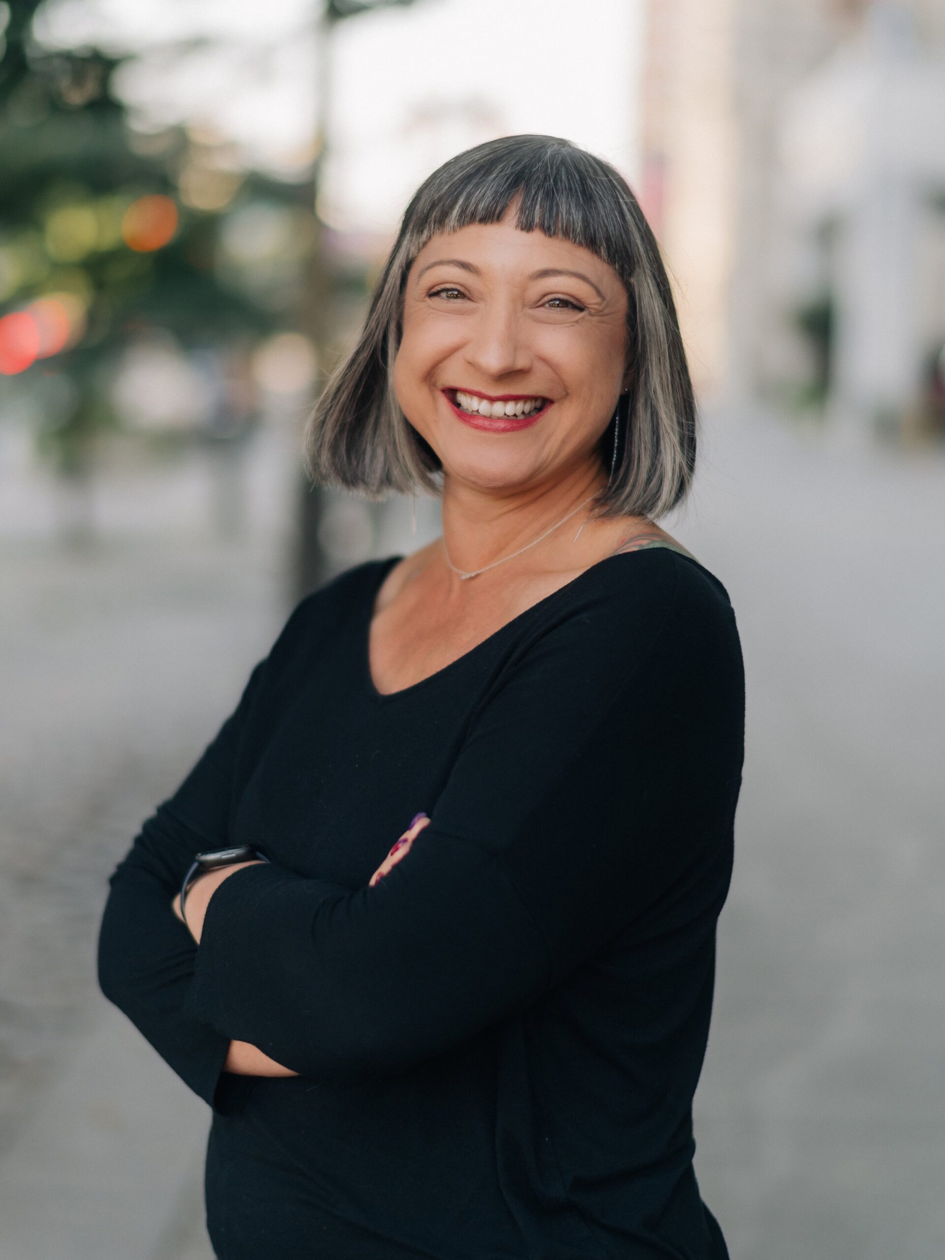 A woman with short gray hair and bangs smiles with arms crossed, standing outdoors on a city street wearing a black top, reflecting the community spirit of Co-operative Housing.