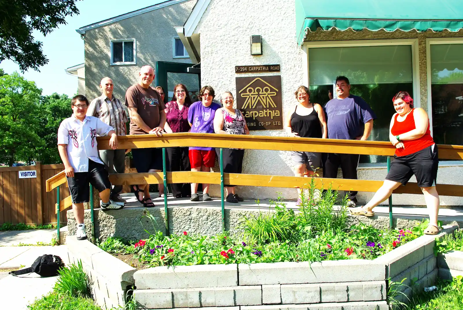 A group of nine people stand and sit outside a building with a "Carpathia" sign, near a flower bed and accessible ramp, on a sunny day—capturing the spirit of co-operative housing supported by CHF Canada.