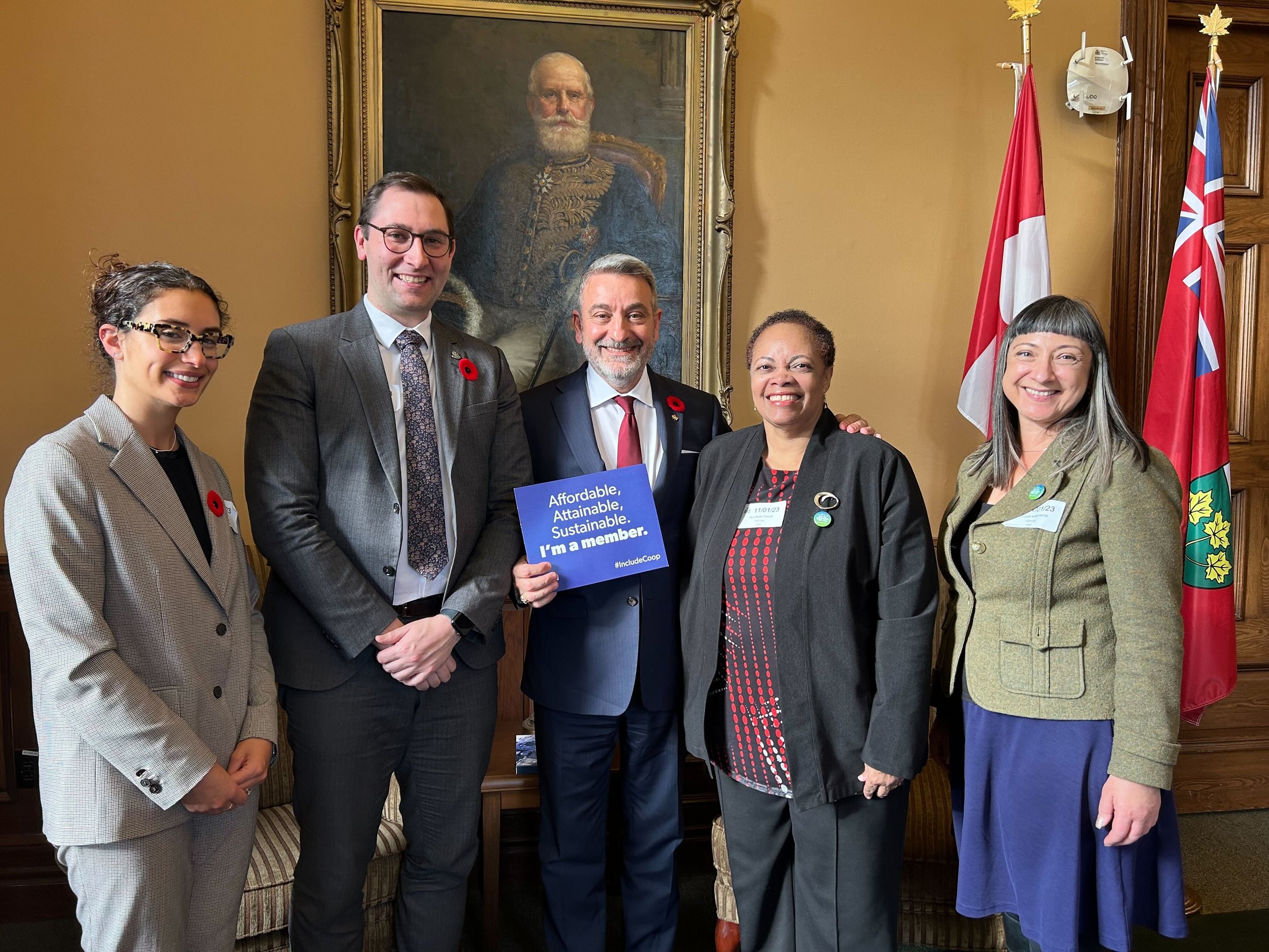 Five people stand together indoors in front of a portrait and flags; the person in the center holds a sign reading, "Affordable. Available. Sustainable. I'm a member." They proudly support co-operative housing with CHF Canada.