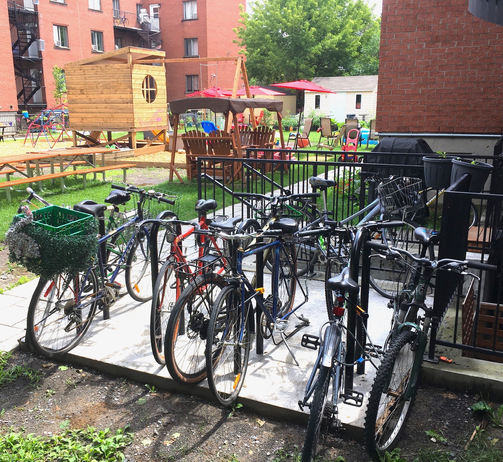 A group of bicycles are parked at a bike rack in front of a playground area with benches and a wooden playhouse, nestled among brick buildings in a Co-operative Housing community.