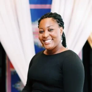 A woman with braided hair, wearing a black long-sleeve top, smiles at the camera with white curtains and a blue patterned background behind her, reflecting the welcoming spirit of Co-operative Housing supported by CHF Canada.
