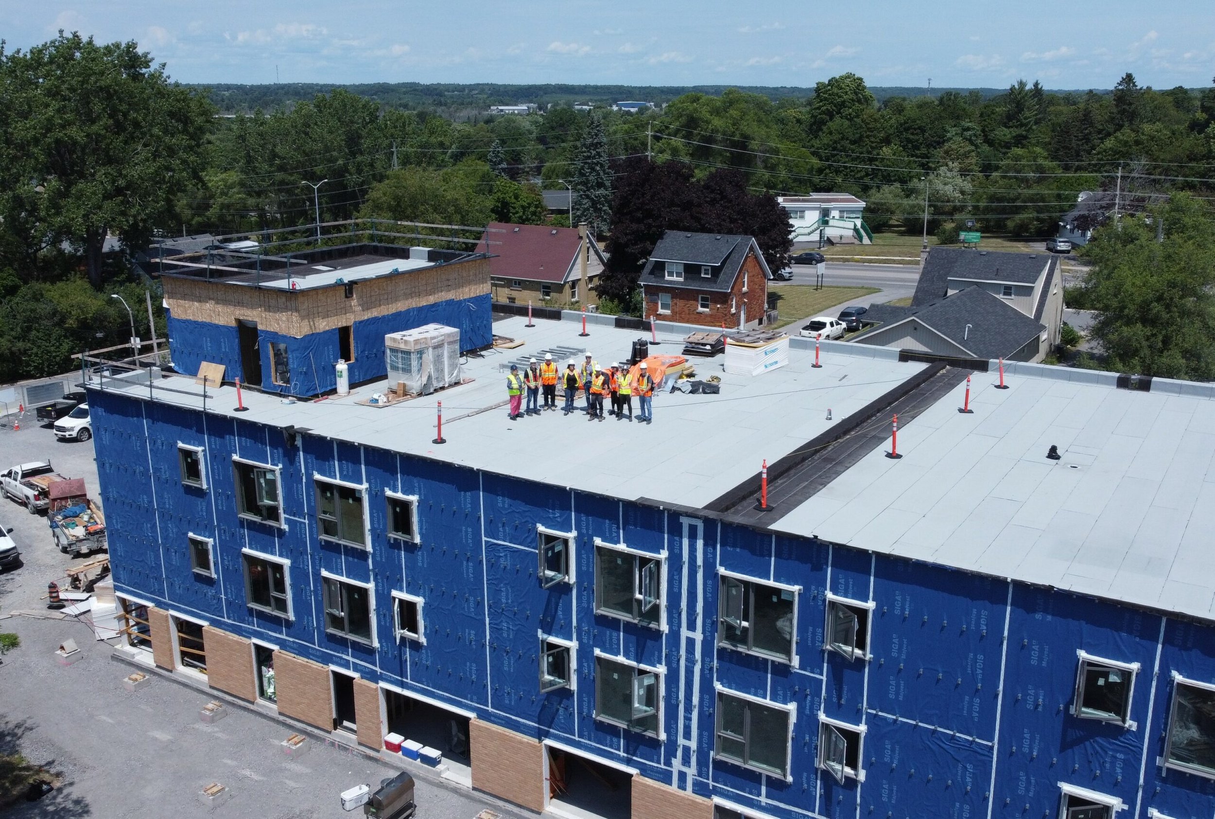 A group of construction workers in safety gear stand on the flat roof of a blue, partially completed co-operative housing building on a clear day.