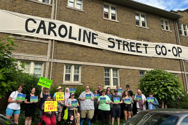 A group of people stand in front of a brick building with a large banner reading "Caroline Street Co-op," holding signs supporting fair housing and tenant rights, celebrating the power of co-operative housing.