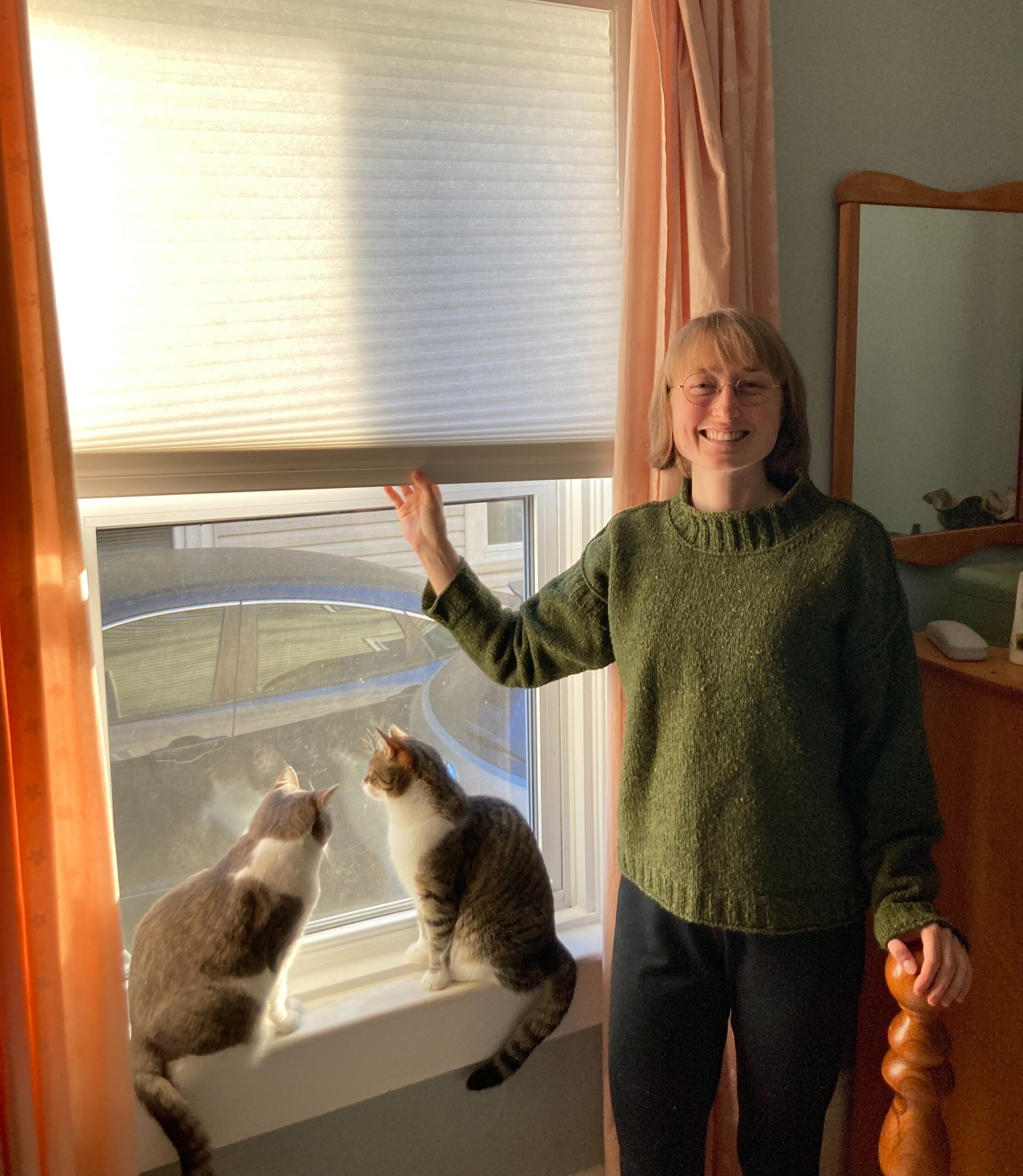 A woman in a green sweater stands by a sunlit window in her co-operative housing unit, with two cats sitting on the windowsill, looking outside.