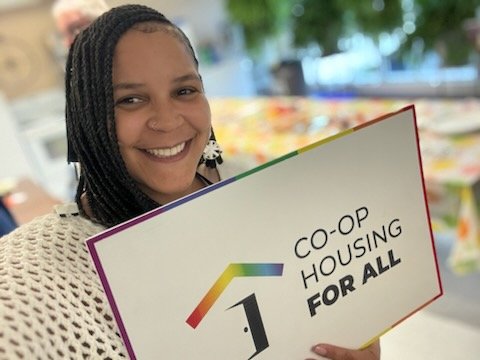 Smiling person holding a sign that reads "Co-op Housing For All" with a rainbow roof graphic above the text, highlighting the importance of Co-operative Housing and support from CHF Canada.