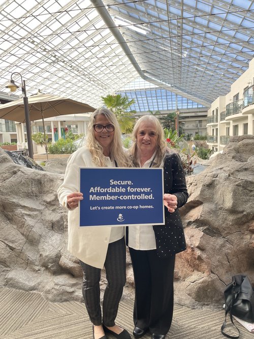 Two women stand indoors, holding a sign reading, "Secure. Affordable forever. Member-controlled. Let's create more co-op homes," promoting Co-operative Housing in Canada with rocks and a glass ceiling in the background.
