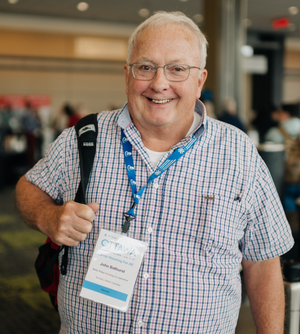 Older man with glasses, wearing a checkered shirt and conference badge, smiles at the camera indoors. He has a backpack strap over one shoulder, a blue lanyard around his neck, and attends a CHF Canada Co-operative Housing event.
