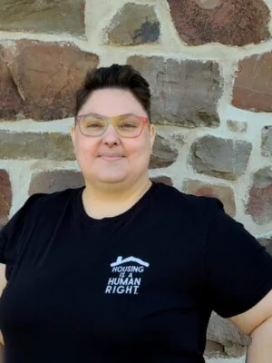 Person with short dark hair and glasses stands in front of a stone wall, wearing a black t-shirt that reads "Housing is a Human Right," showing support for co-operative housing and organizations like CHF Canada.
