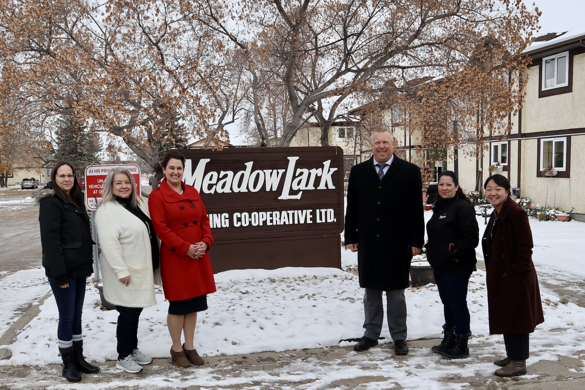 Six people stand in front of a large "Meadowlark Housing Cooperative Ltd." sign outside, with snow on the ground and trees in the background, highlighting the strong sense of community found in co-operative housing supported by CHF Canada.