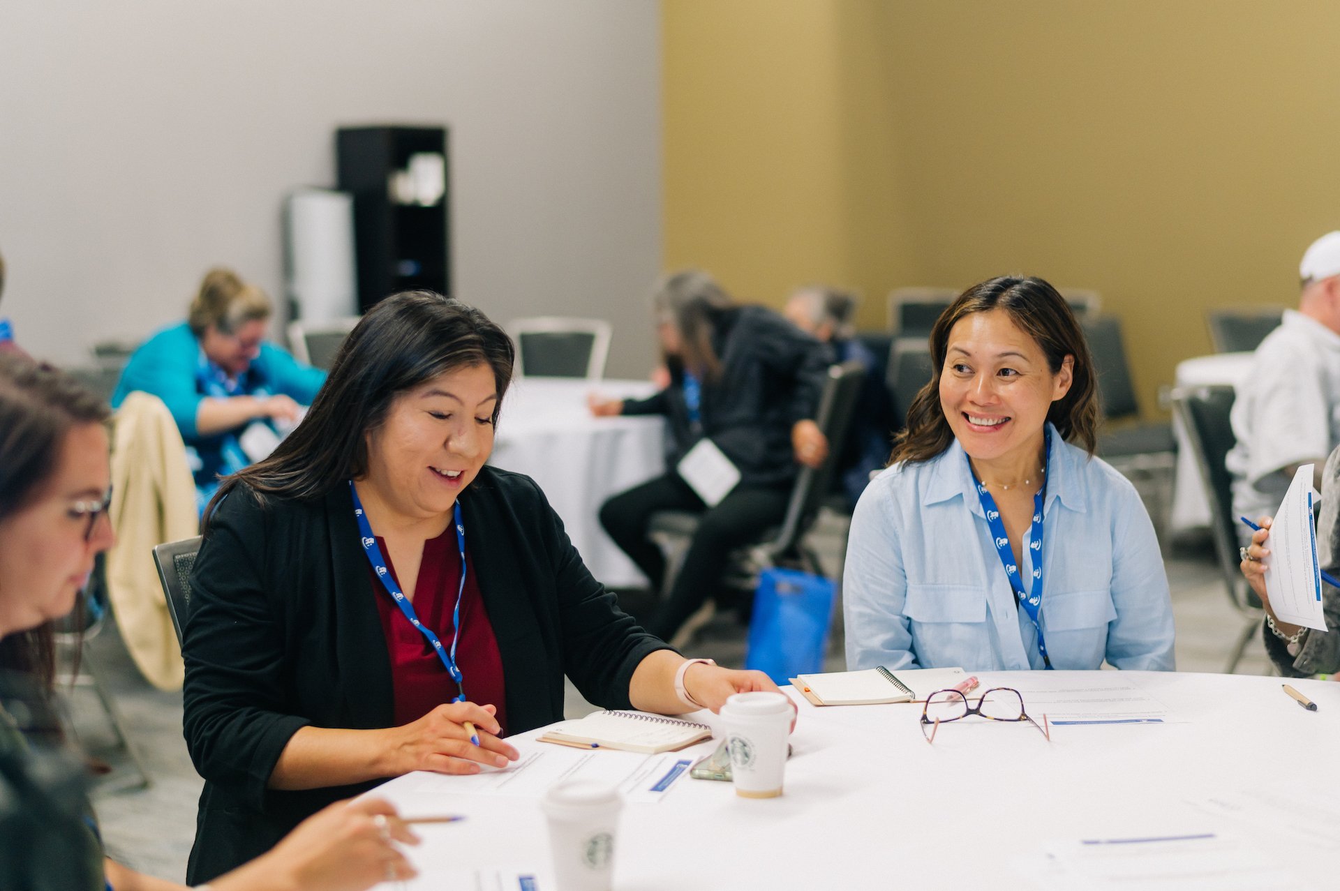 Four people sit around a table in a conference room, writing on papers and talking. Two women with blue lanyards smile, engaged in a Co-operative Housing discussion. Other attendees are blurred in the background at this CHF Canada event.