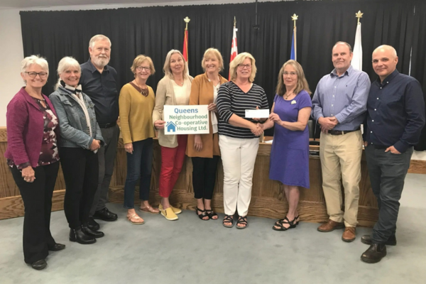 Ten adults stand indoors, some holding a sign reading "Queens Neighborhood Co-operative Housing Ltd" and an envelope, in front of a table with flags—highlighting their commitment to Co-operative Housing and the values supported by CHF Canada.