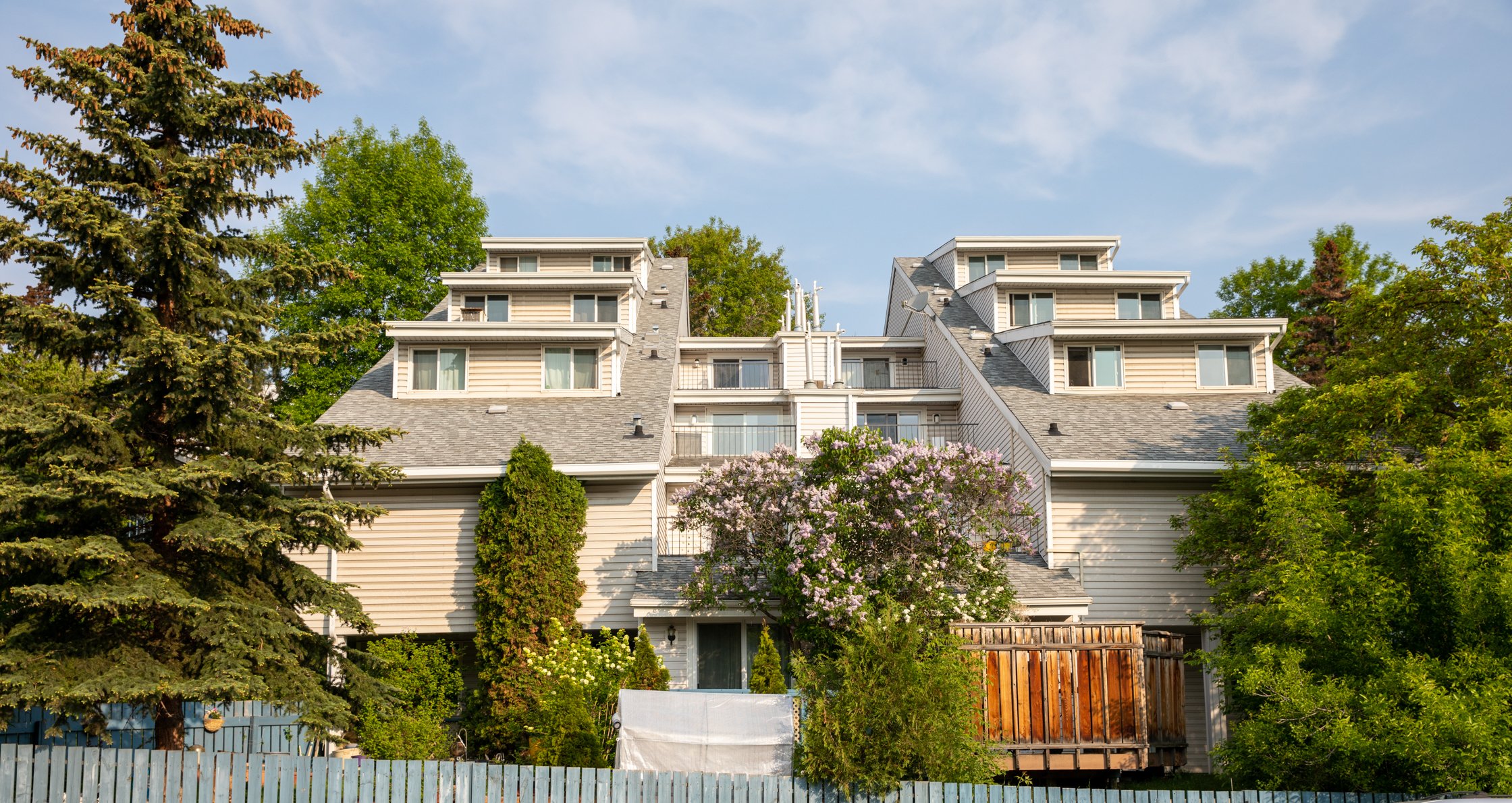 Three-story townhouse building with light-colored siding, multiple peaked roofs, and several windows—an inviting example of co-operative housing—surrounded by trees and a blue wooden fence in the foreground.