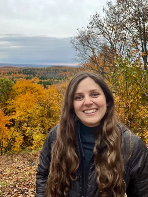 A woman with long brown hair and a black jacket smiles in front of a forest with autumn foliage and overcast sky, capturing the spirit of co-operative housing and community living supported by CHF Canada.