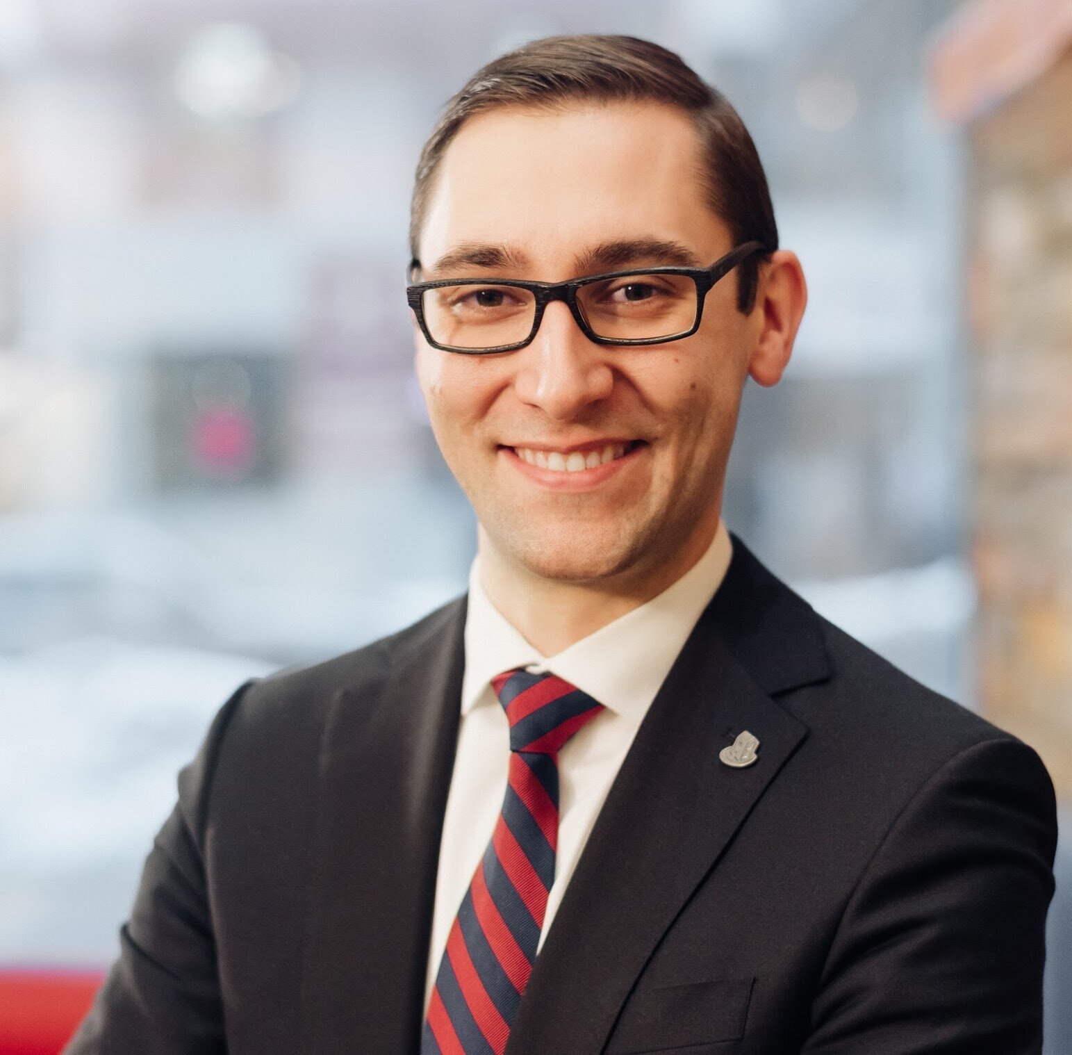 A man in a suit, tie, and glasses stands indoors with arms crossed, smiling at the camera—representing Co-operative Housing and CHF Canada. The background is blurred.
