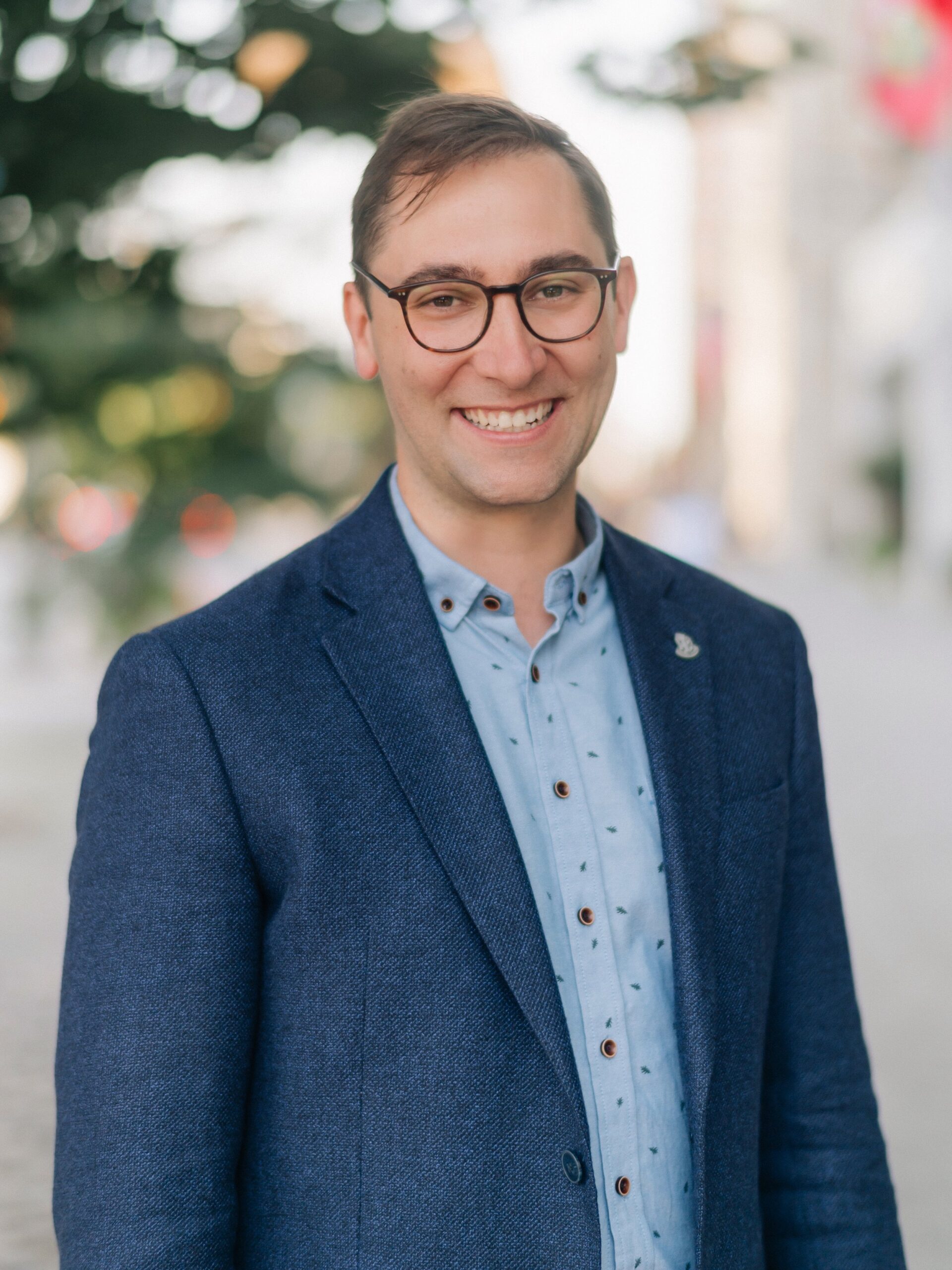 A man wearing glasses, a blue blazer, and a light blue shirt stands outdoors, smiling at the camera with a blurred background of trees and buildings—reflecting the spirit of co-operative housing supported by CHF Canada.