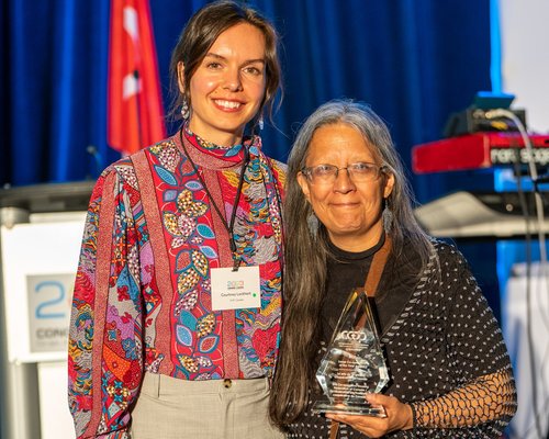 Two women pose together indoors; one holds a clear glass award. Both are smiling, with CHF Canada conference materials and a podium in the background, highlighting their dedication to Co-operative Housing.