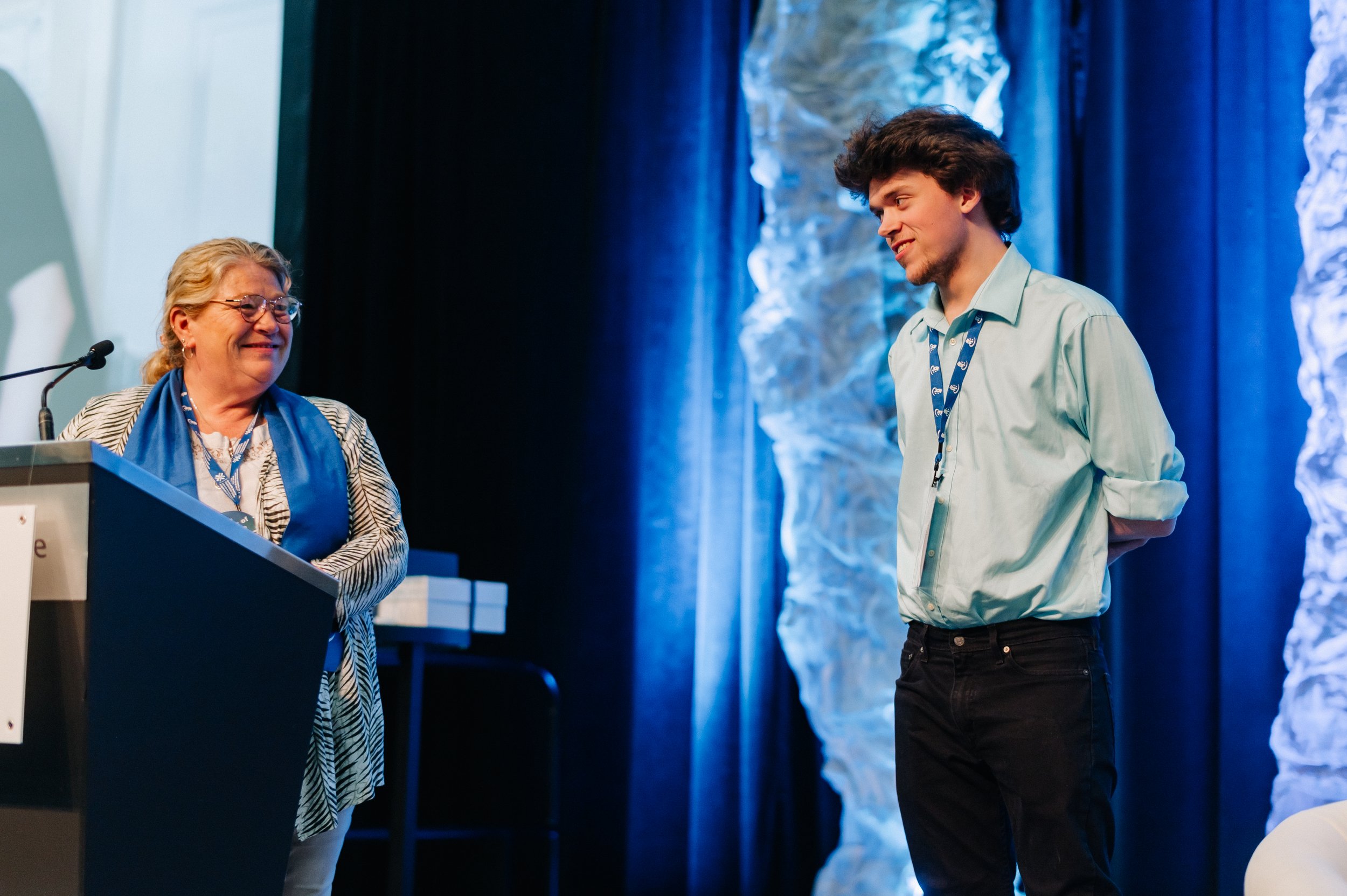A woman stands at a podium speaking to a man wearing a blue shirt and lanyard on stage, with blue lighting and abstract decor in the background at a CHF Canada co-operative housing event.