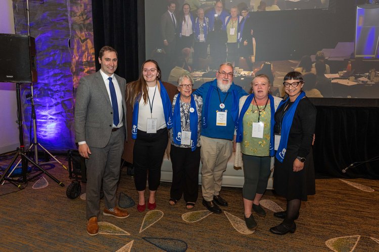Six people pose together indoors, all wearing blue scarves, with a large screen showing a similar group photo in the background—showcasing their pride in Co-operative Housing and connection to CHF Canada.