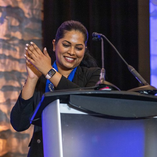 A woman stands at a podium, smiling and clapping, with microphones in front of her and a textured wall in the background at a CHF Canada co-operative housing event.