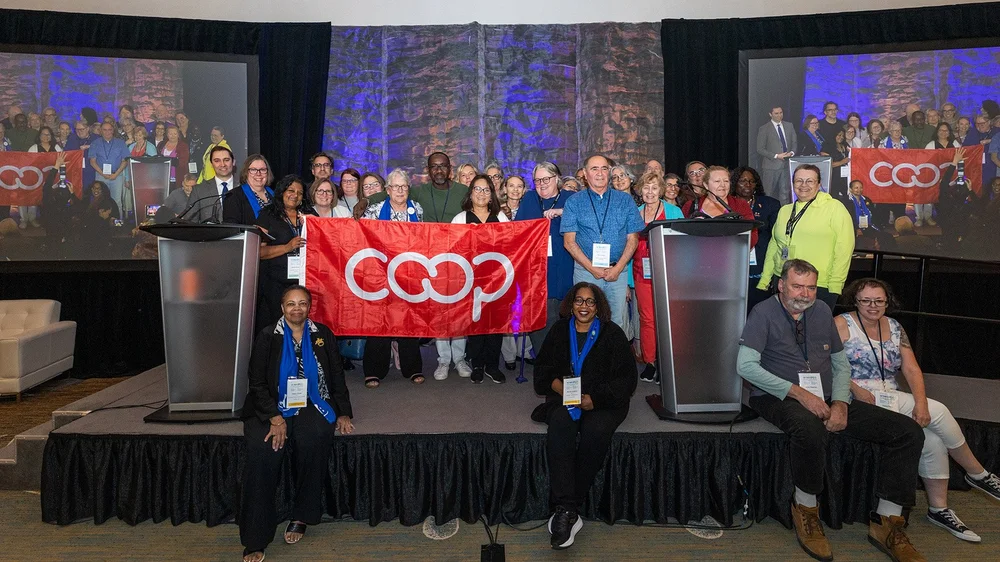 A group of people stands and sits on a stage holding a red COOP flag, representing Co-operative Housing, with two podiums and large screens displaying their image in the background at a CHF Canada event.