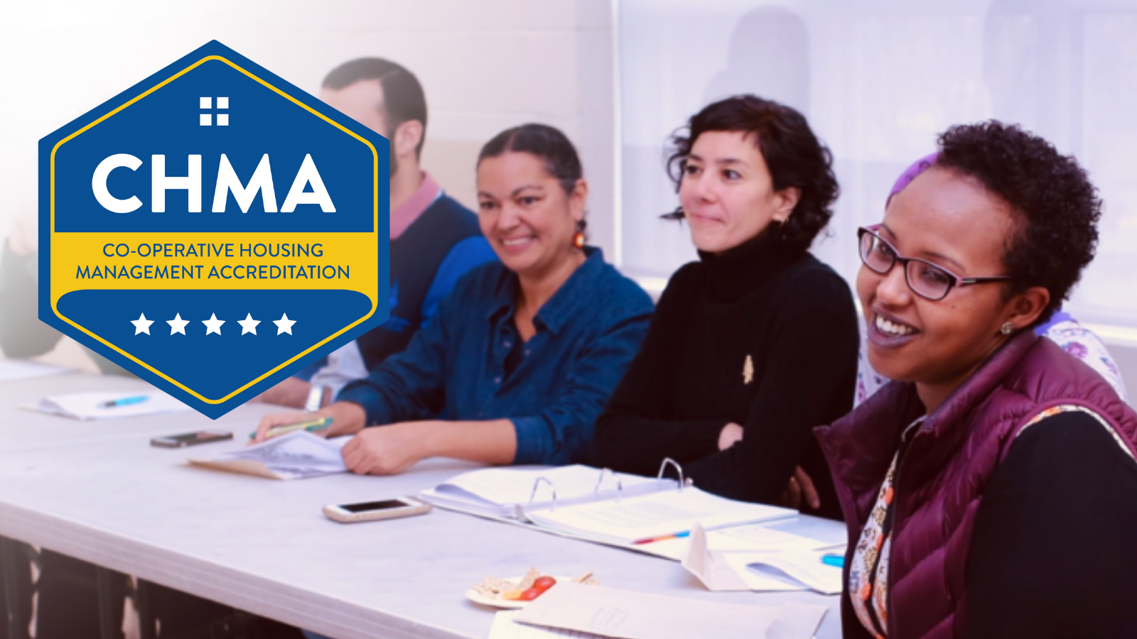 Four people sit at a table with notebooks, listening and smiling during a meeting about co-operative housing. A large CHMA (Co-operative Housing Management Accreditation) badge is overlaid, highlighting CHF Canada’s commitment to excellence in housing.