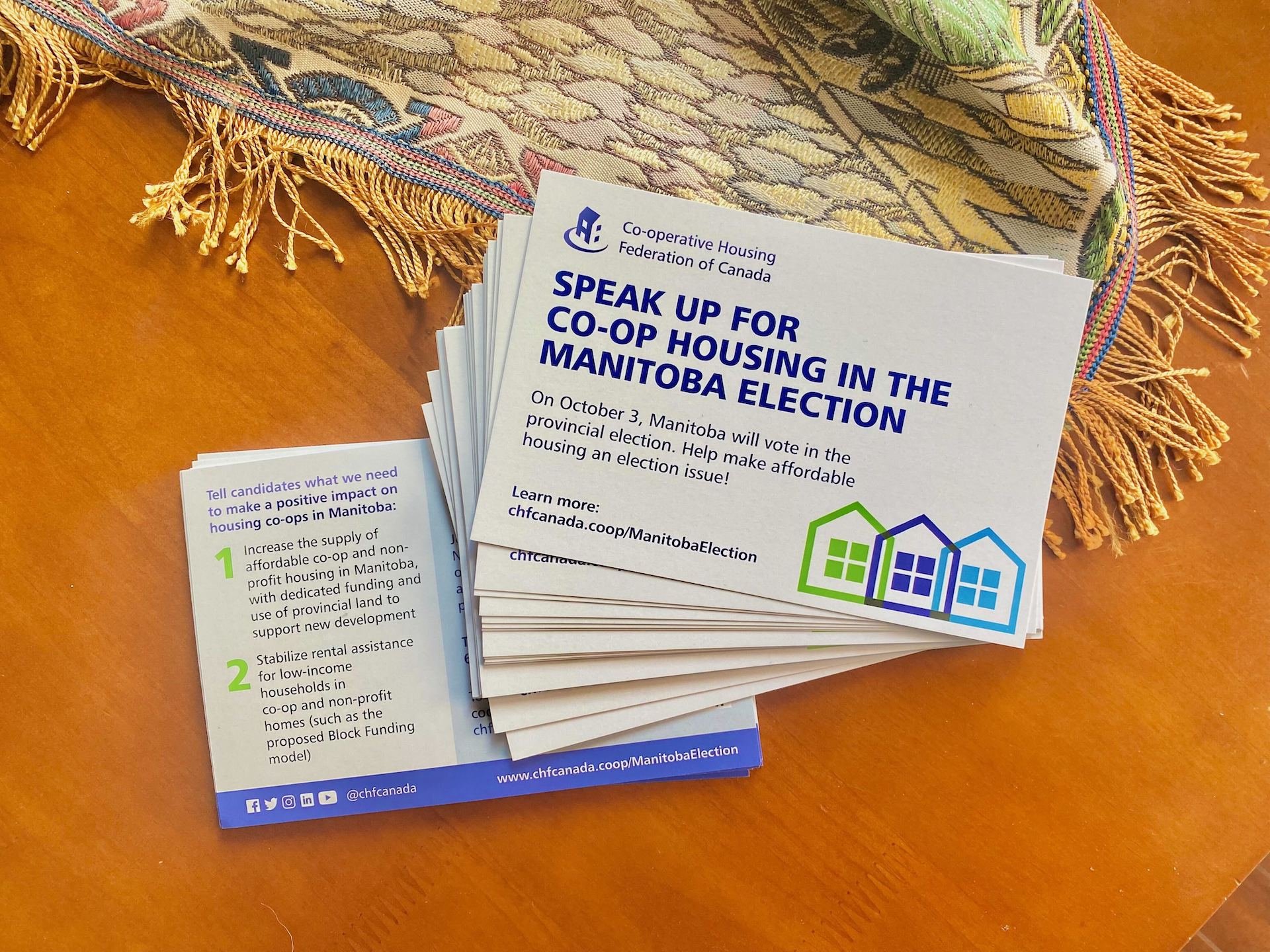 A stack of postcards on a wooden table urging voters to support co-operative housing in the Manitoba election, featuring information, colorful house icons, and highlighting CHF Canada’s role in advocating for better housing solutions.