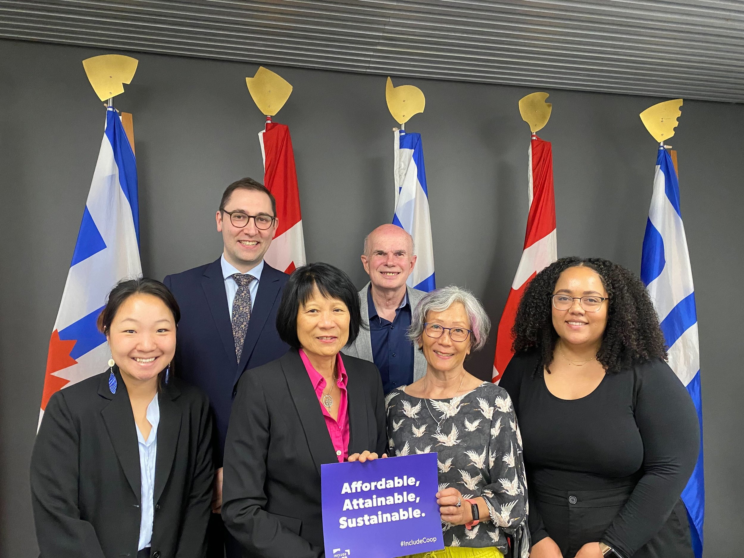 Six people pose indoors in front of five flags; one person holds a sign that says, "Affordable, Attainable, Sustainable," highlighting the importance of co-operative housing and support from organizations like CHF Canada.