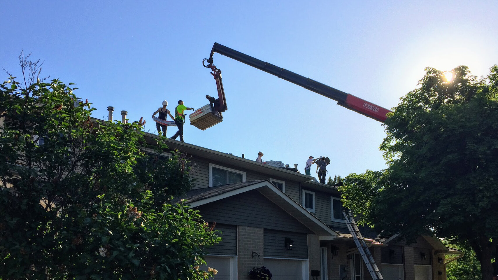 Workers on a house roof receive building materials lifted by a crane, highlighting the teamwork found in co-operative housing. Lush trees and a clear sky provide the backdrop, reflecting CHF Canada’s commitment to quality housing.