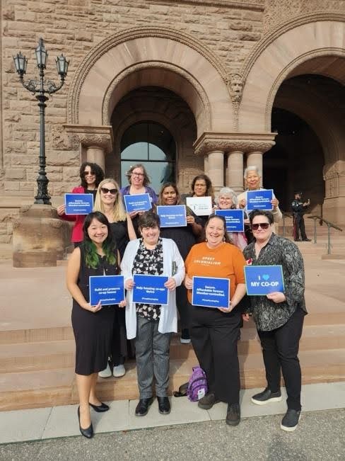 A group of people stand on stone steps outside a historic building, holding blue and orange signs supporting Co-operative Housing and CHF Canada.